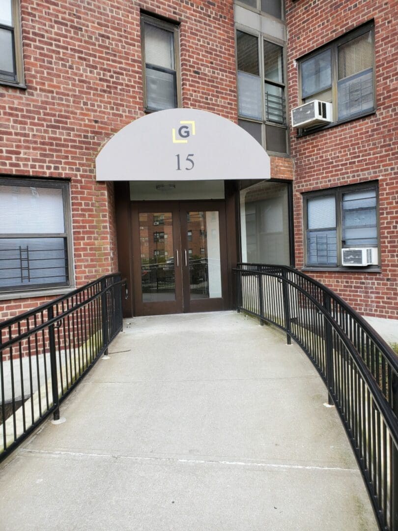Entrance to a brick apartment building numbered 15, featuring a glass door under a beige awning with the letter 