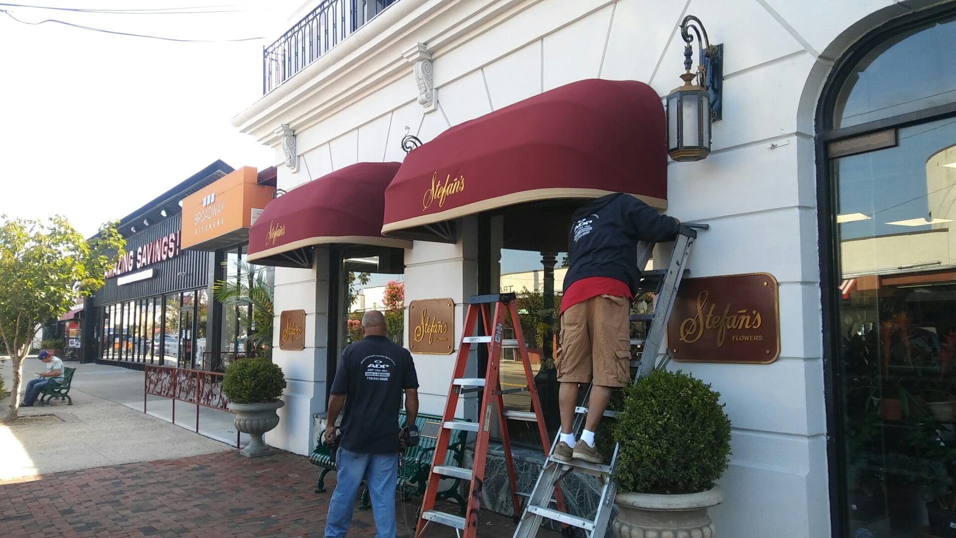 Two workers on ladders installing red awnings on a storefront with the name 