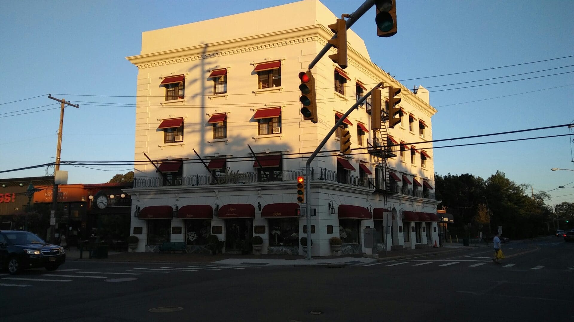 A three-story, white building with red awnings and ground-floor businesses, including the ADP USA Solutions Gallery, located at a sunlit street intersection with traffic lights and a pedestrian crossing.