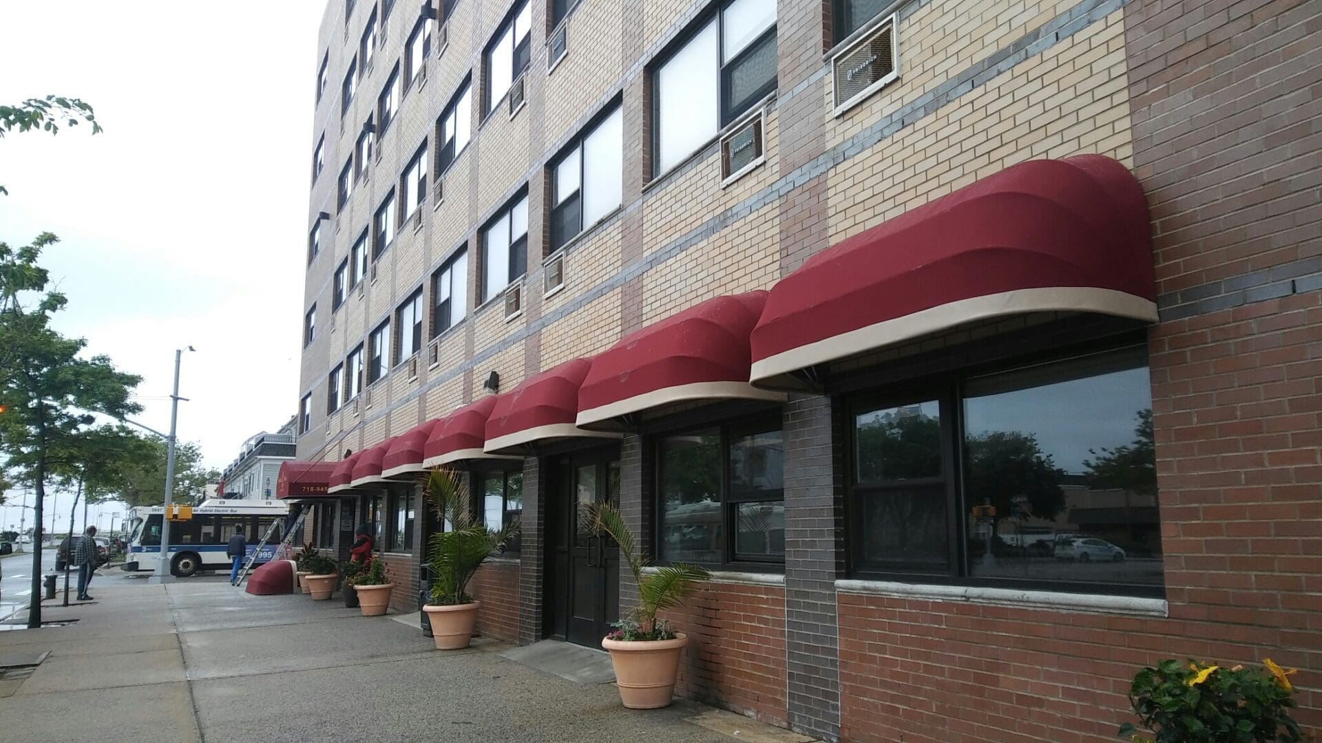 Brown brick building with red awnings over windows and doorway, lined with potted plants on a cloudy day, serving as the ADP USA Solutions Gallery.