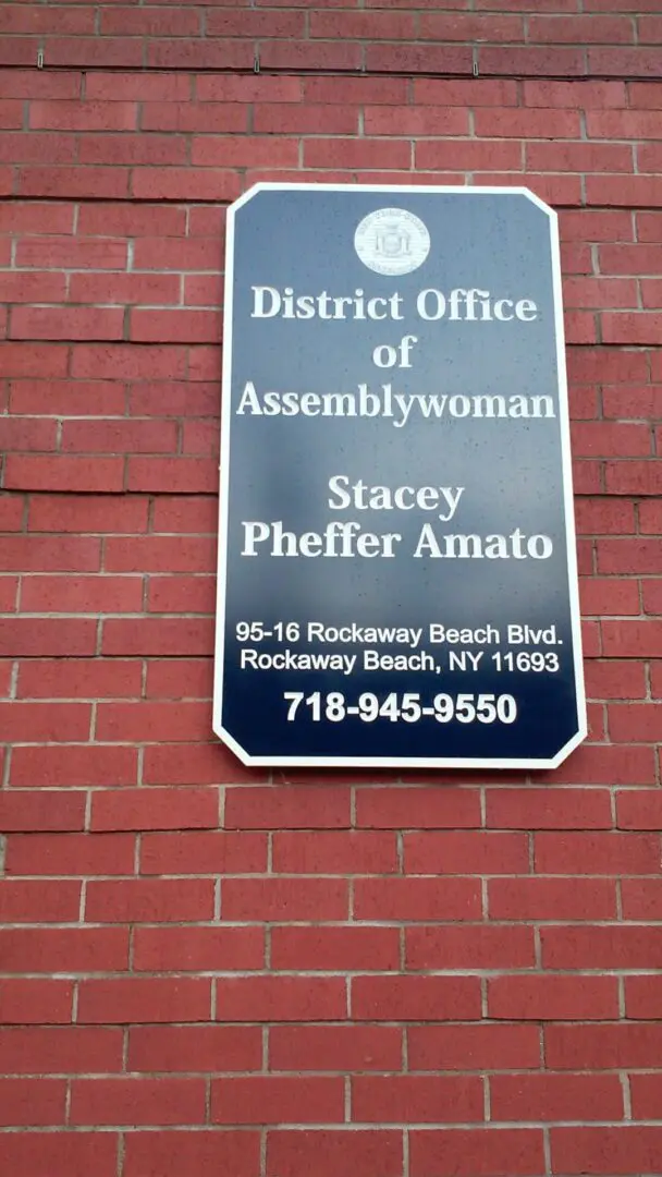 Sign on a brick wall for the district office of Assemblywoman Stacey Pheffer Amato located at Rockaway Beach, NY, displaying contact details near the ADP USA Solutions Gallery.