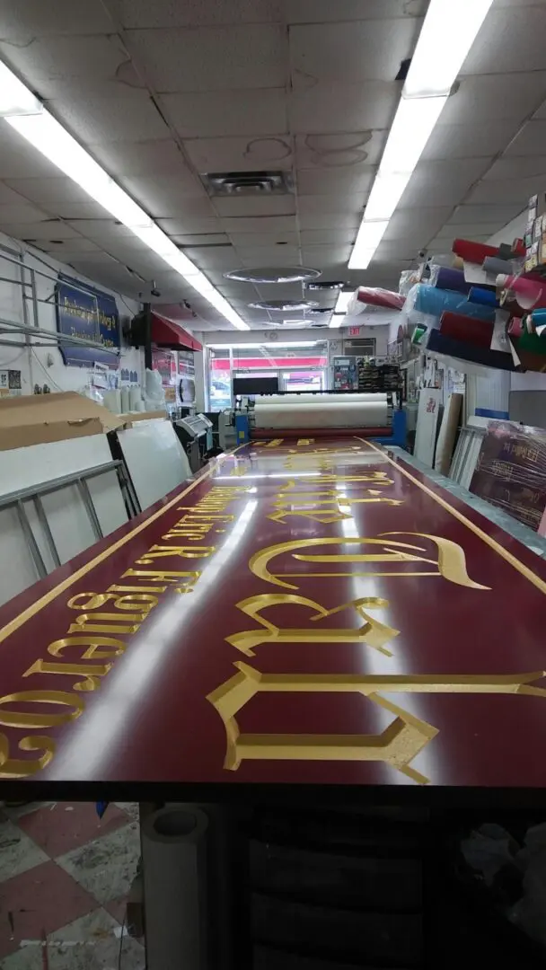 Interior of a print shop with a long, freshly printed red and gold banner on a table, surrounded by rolls of paper and signage materials at the ADP USA Solutions Gallery.