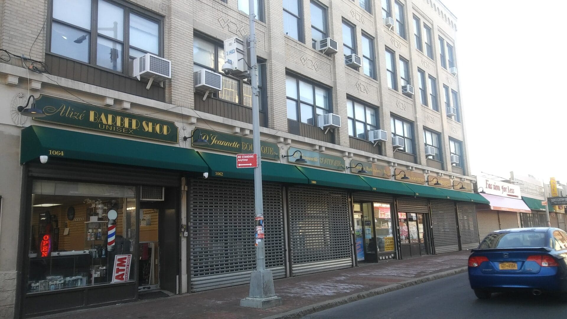 Street view of a row of storefronts with closed security shutters and a passing blue car on a sunny day, featuring an ADP USA Solutions Gallery.