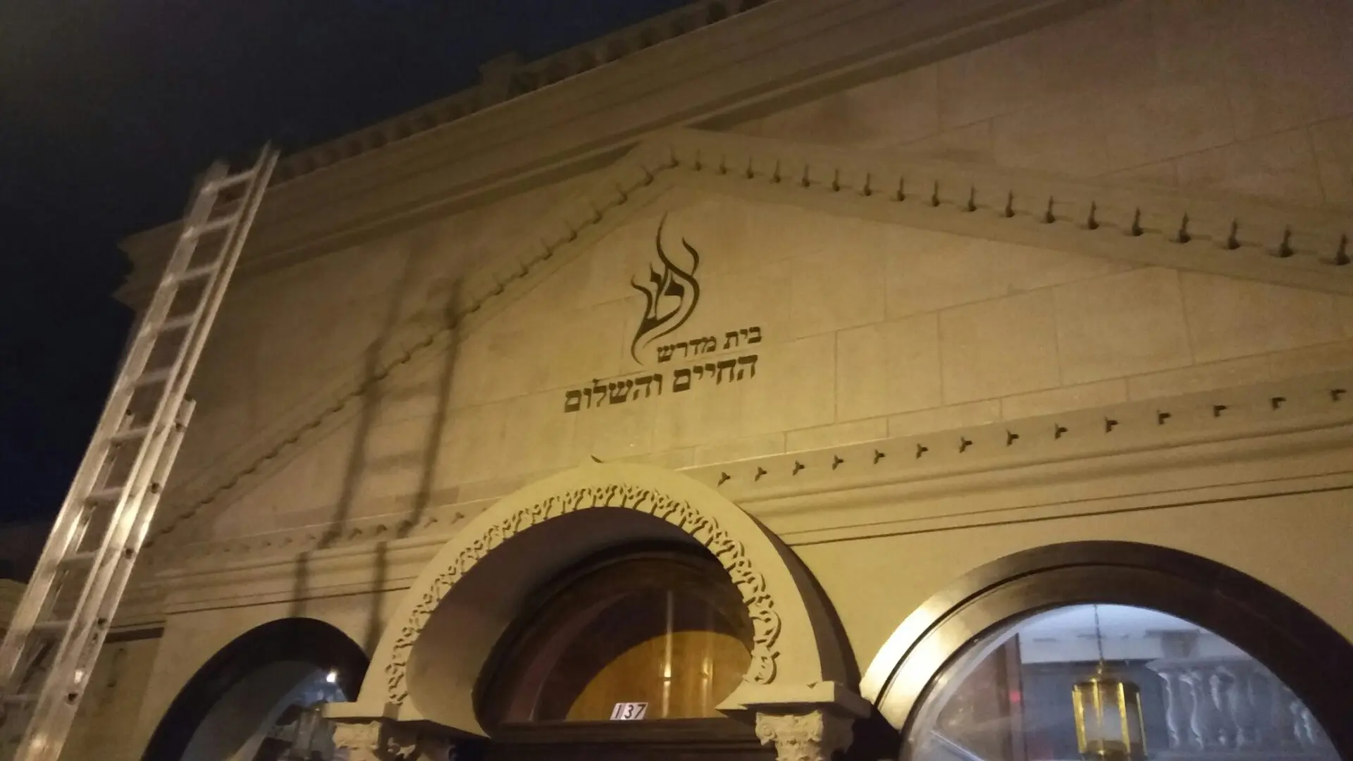 Night view of the ADP USA Solutions Gallery facade with Hebrew text, ornate arches, and a decorative flame symbol, illuminated by soft lighting.