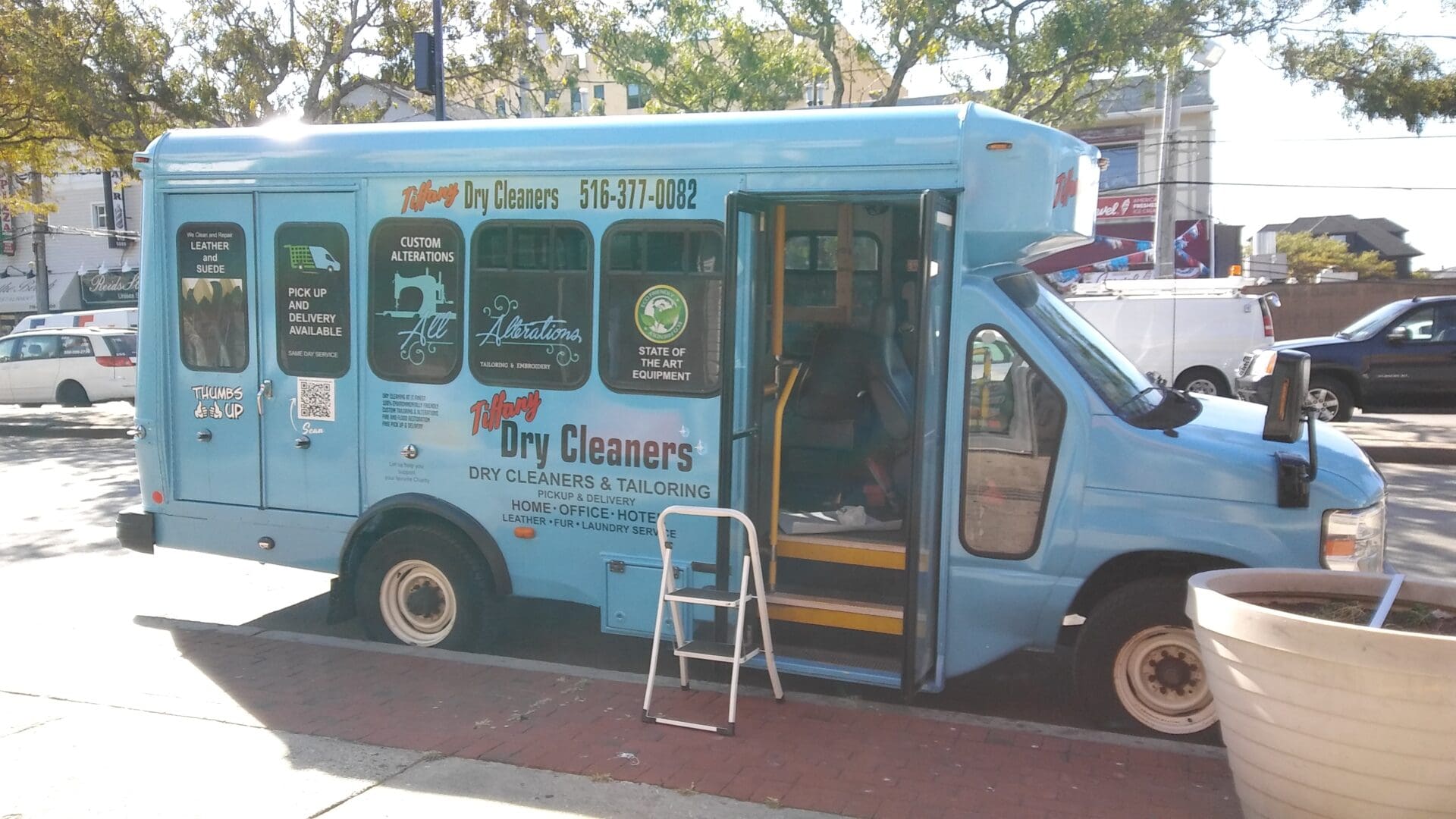A blue mobile dry cleaning van parked on a street with its side door open, displaying various ADP USA Solutions Gallery advertisements.
