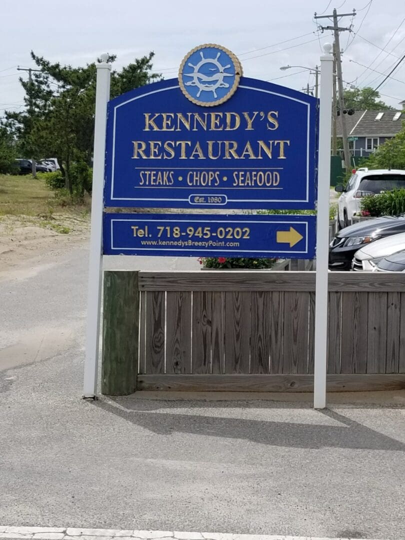 Blue and white sign for Kennedy's restaurant advertising steaks, chops, and seafood, with contact information, set against a backdrop of a parking lot and greenery at ADP USA Solutions Gallery.