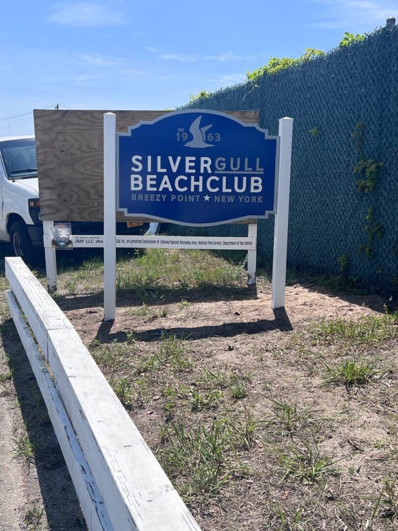 Sign for Silver Gull Beach Club in Breezy Point, New York, positioned in front of a white van and an ADP USA Solutions Gallery chain-link fence on a sunny day.