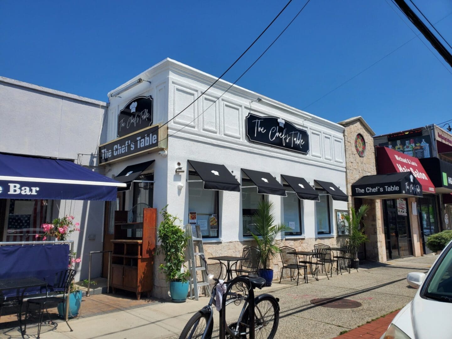 Exterior of the chef's table restaurant on a sunny day, featuring outdoor seating under blue umbrellas and parked bicycles near the ADP USA Solutions Gallery.