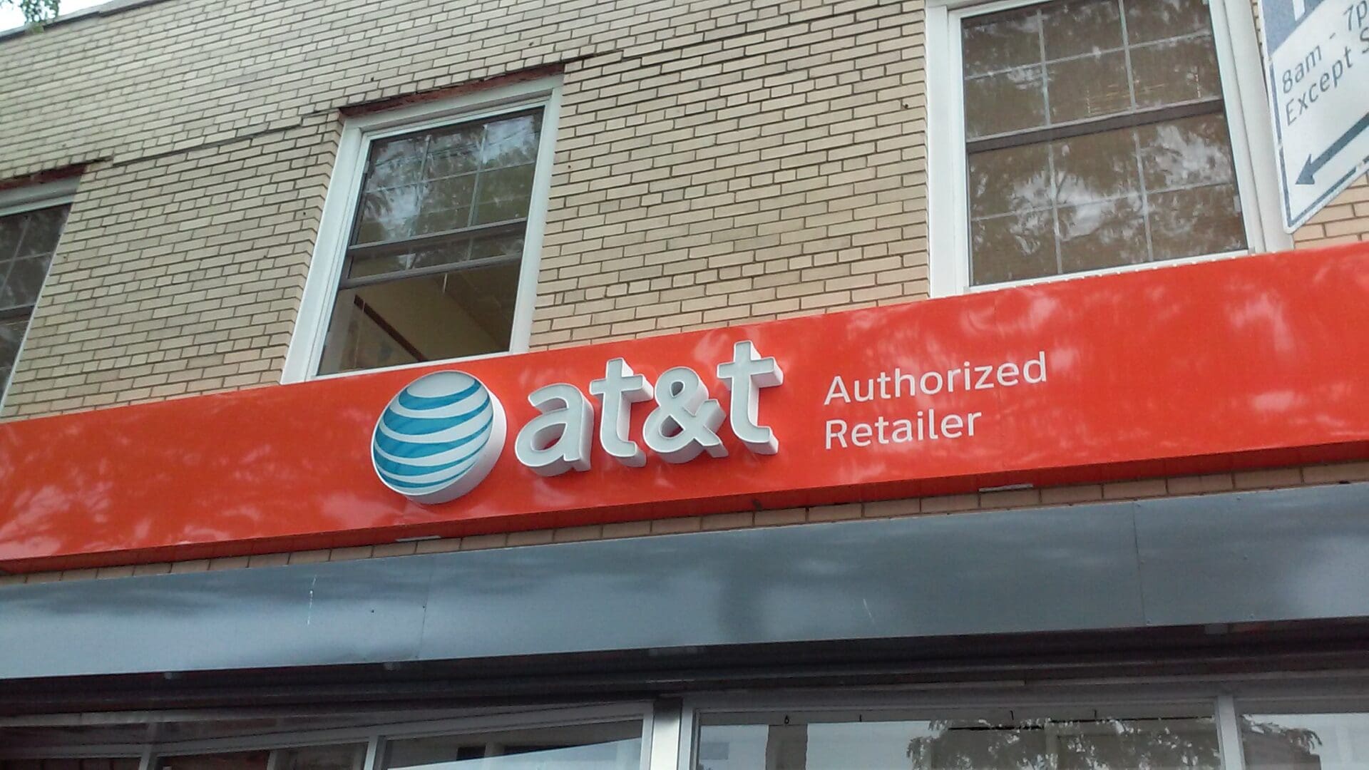 Red and white AT&T authorized retailer sign on a building facade with windows and trees reflected in the windows, near the ADP USA Solutions Gallery.